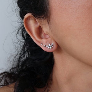 May include: A close-up of a person's ear with two silver earrings. The earring on the top is a small bee design. The earring on the bottom is a small silver ball.