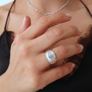 Peut inclure: Une large bague en m&eacute;tal argent&eacute; pr&eacute;sentant une texture martel&eacute;e et un motif d'&oelig;il grav&eacute; en son centre. Le design comprend une pupille d&eacute;taill&eacute;e et des lignes rayonnantes entourant la forme de l'&oelig;il, cr&eacute;ant un motif visuel sur la surface.