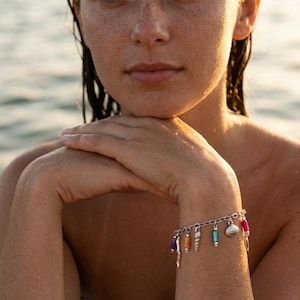 May include: A silver chain charm bracelet featuring a variety of dangling elements, including a small silver seashell, a textured silver bar, and cylindrical beads in vibrant shades of purple, orange, turquoise, and bright pink, secured with a clasp.
