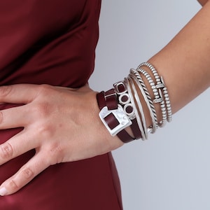 Op de afbeelding: Een stapel zilveren armbanden met een bruin leren armband met een zilveren gesp. Het leren armband heeft een zilveren bedel met drie cirkels.