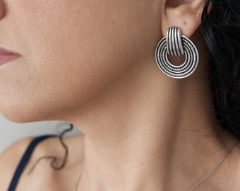 Puede incluir: Un pendiente de metal plateado con un diseño circular de anillos concéntricos texturizados y un enganche superior vertical acanalado. El pendiente tiene un acabado mate y un aspecto superpuesto que muestra una combinación de detalles lisos y con cuentas.