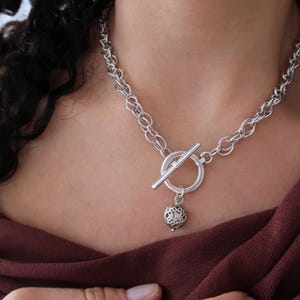 May include: A silver chain necklace with a toggle clasp and a dangling ball charm. The charm has a detailed, intricate design.