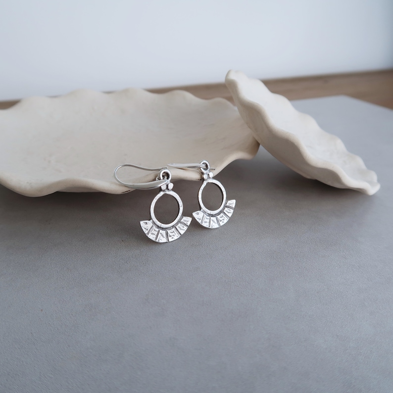 May include: A pair of silver earrings with a circle design and a fan-shaped bottom. The earrings are hanging from a white surface.