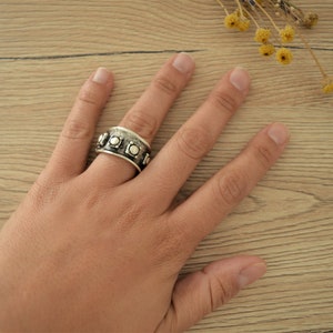 May include: A silver ring with a textured band and round silver accents. The ring is worn on a person's finger.