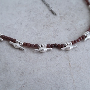 May include: A brown gemstone beaded necklace with silver eye charms. The necklace is made with small, round beads and has three silver charms with an eye design.