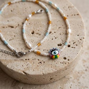 May include: A beaded necklace featuring small white seed beads, interspersed with light blue and amber-colored beads, irregular white pearls, and a central round charm depicting a stylized eye with a rainbow-colored base and a silver metal clasp.