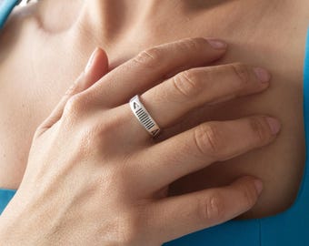 May include: A silver ring with a geometric design. The ring features a series of vertical lines and a triangular shape. The ring is worn on a finger. The background is a blue top.