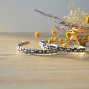 May include: Two silver bracelets with a decorative pattern on a wooden surface. The bracelets are stacked on top of each other.