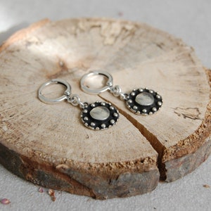May include: A pair of silver hoop earrings with a round, silver pendant featuring a textured, circular design with a raised center. The pendant is surrounded by a row of small, silver beads.