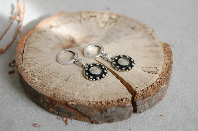 May include: A pair of silver hoop earrings with a round, silver pendant featuring a textured, circular design with a raised center. The pendant is surrounded by a row of small, silver beads.