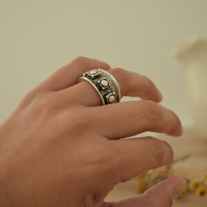May include: A silver ring with a wide band and multiple round accents. The ring is worn on a person's finger.