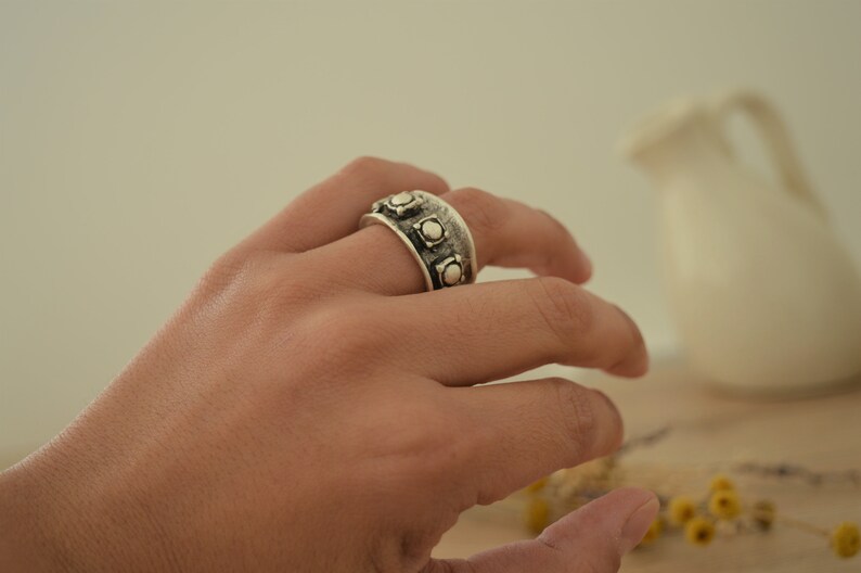 May include: A silver ring with a wide band and multiple round accents. The ring is worn on a person's finger.