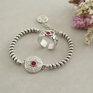Puede incluir: Un brazalete de plata con un diseño trenzado y un anillo de plata con una piedra roja. El brazalete tiene un dije con una piedra roja y un diseño floral.