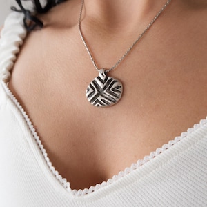 Op de afbeelding: Een zilveren ketting met een hanger. De ronde, getextureerde hanger heeft een geometrisch patroon. De ketting heeft een fijne zilveren ketting. De ketting wordt gedragen over een witte top.