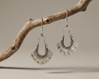 May include: A pair of silver-toned dangle earrings featuring a fan-shaped, textured metal base with a central open cutout. The design includes a beaded border along the inner edge and a simple hook wire for attachment, showcasing a metallic finish.