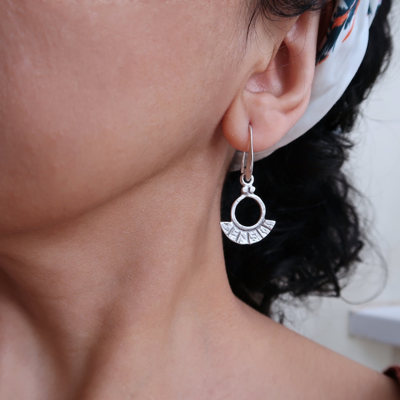 May include: Silver hoop earrings with a textured, fan-shaped design. The earrings are dangling from the earlobe.