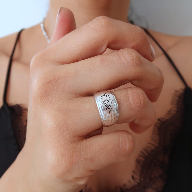 Peut inclure: Une large bague en m&eacute;tal argent&eacute; avec une texture martel&eacute;e et un motif d'&oelig;il grav&eacute; au centre, accompagn&eacute; de lignes rayonnantes. La surface m&eacute;tallique pr&eacute;sente une finition mate et le motif est centr&eacute; sur la circonf&eacute;rence ext&eacute;rieure.