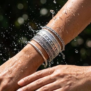 Könnte beinhalten: Ein Stapel aus vier silberfarbenen Metallarmbändern an einem Unterarm. Das Set besteht aus einem dünnen Drahtarmreif mit texturiertem Mittelband, einer breiten Manschette mit filigranen Mustern, einem Band mit geprägten Punkten und einem geflochtenen Kettenarmband.