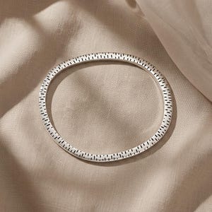 Puede incluir: Una pulsera rígida circular de plata con una superficie texturizada que presenta un patrón repetitivo de pequeñas rayas negras verticales y finas grabadas a lo largo de toda la circunferencia exterior de la banda metálica, creando un efecto visual.