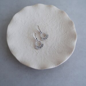 May include: A pair of silver earrings with a circle and fan design. The earrings are on a white ceramic dish.