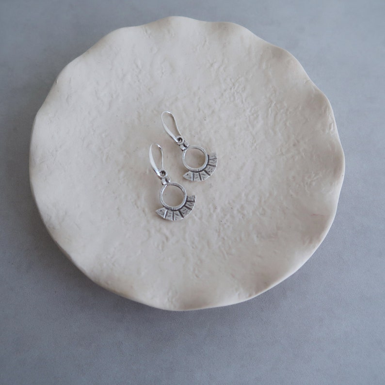 May include: A pair of silver earrings with a circle and fan design. The earrings are on a white ceramic dish.