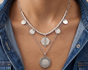 May include: A layered silver-tone necklace featuring a top strand with small round beads and four small circular charms. The lower strand includes a central sunburst medallion pendant and a bottom circular pendant with a textured, dimpled surface.