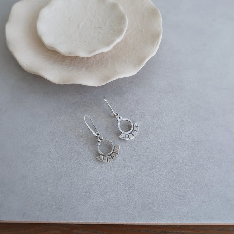 May include: A pair of silver earrings with a circle design and a fan-shaped detail. The earrings are on a gray surface.