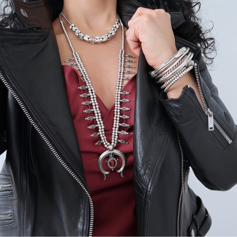 May include: A woman wearing a black leather jacket with a silver necklace featuring a large pendant. The necklace has a layered design with multiple strands of beads and a silver charm.