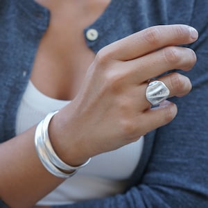 May include: A silver ring with a textured, organic shape. The ring is worn on the middle finger of a person's hand. The person is also wearing a silver bangle bracelet on their wrist.