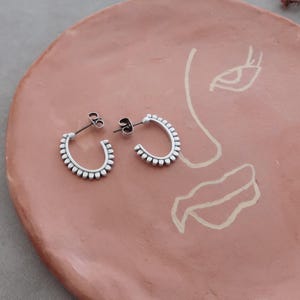 May include: A pair of silver hoop earrings with a beaded design. The earrings are on a terracotta-colored surface.