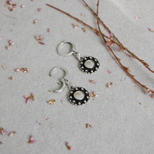 May include: A pair of silver hoop earrings with a small, round, black disc pendant. The disc has a textured surface and a raised edge.