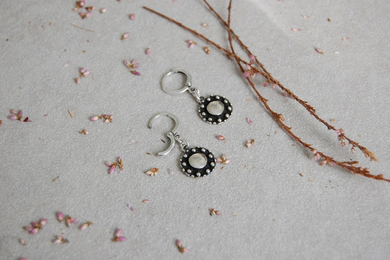 May include: A pair of silver hoop earrings with a small, round, black disc pendant. The disc has a textured surface and a raised edge.