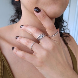 May include: A hand wearing multiple silver rings. The rings are stacked on the middle finger and a single ring is on the ring finger. The rings are all thin and delicate.
