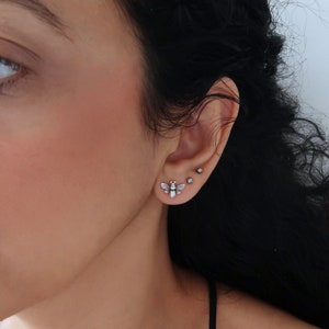 May include: A close-up of a person's ear with a silver bee stud earring. The earring is small and delicate, and it is set against a backdrop of dark hair.
