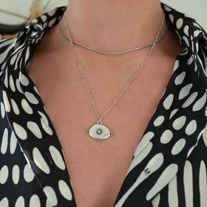 May include: A silver necklace with a pendant shaped like an eye with a small circle in the centre. The necklace is layered over a thin silver chain.