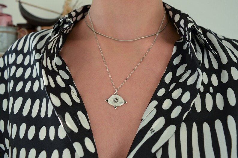 May include: A silver necklace with a pendant shaped like an eye with a small circle in the centre. The necklace is layered over a thin silver chain.
