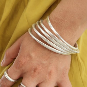 Pode incluir: Um conjunto de cinco pulseiras de prata empilhadas no pulso de uma pessoa. As pulseiras são lisas e brilhantes, e todas têm a mesma largura.