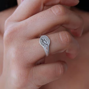 Puede incluir: Un anillo de plata con un diseño en forma de corazón que presenta un pájaro en su interior. El anillo se lleva en un dedo.