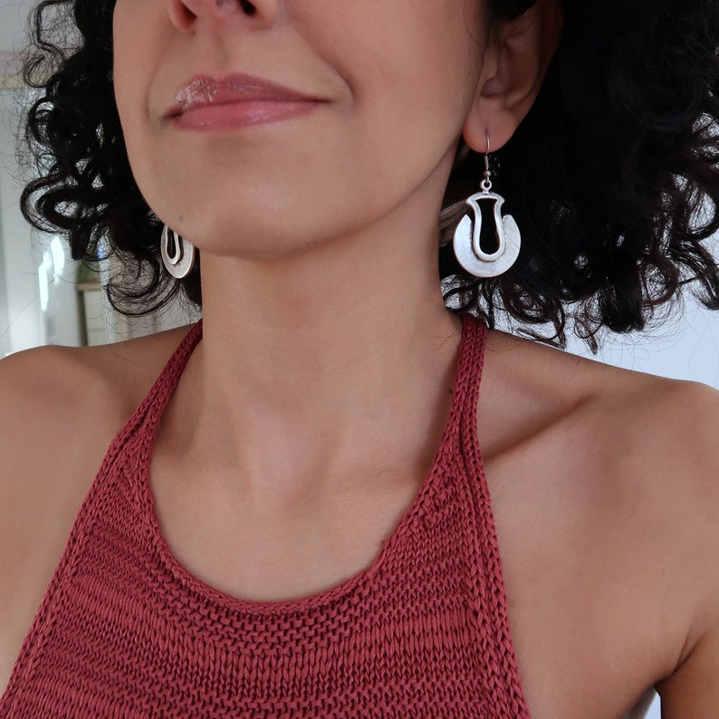 May include: A woman wearing a red knitted halter top with silver hoop earrings. The earrings have a unique geometric design.