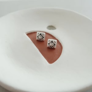 May include: Two silver square stud earrings with a textured design on a white ceramic dish with a brown crescent moon shape.
