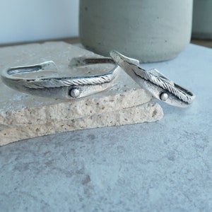 Puede incluir: Dos pulseras de plata con un diseño de cuerda y un pequeño acento de cuentas. Las pulseras están apiladas una encima de la otra sobre una superficie beige claro.