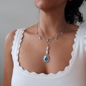 May include: A silver necklace with a blue gemstone pendant and smaller blue gemstone accents. The pendant features a detailed design with a circular frame and a delicate chain.