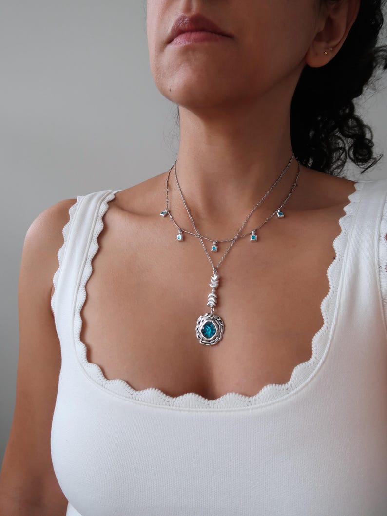 May include: A silver necklace with a blue gemstone pendant and smaller blue gemstone accents. The pendant features a detailed design with a circular frame and a delicate chain.