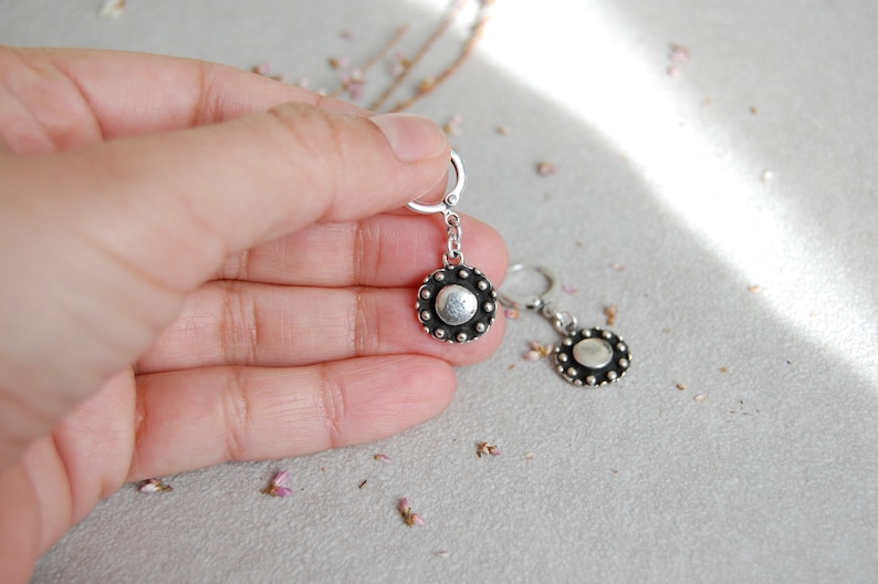 May include: A pair of silver earrings with a round, textured disc pendant. The discs have a silver center and a black outer ring with small silver dots.