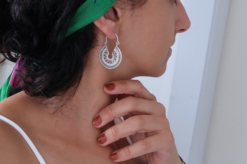 Puede incluir: Pendientes de aro plateados con un dise&ntilde;o calado detallado. Los pendientes tienen forma de semic&iacute;rculo con patrones intrincados, creando una apariencia delicada y ornamental. El dise&ntilde;o recuerda motivos florales o geom&eacute;tricos.