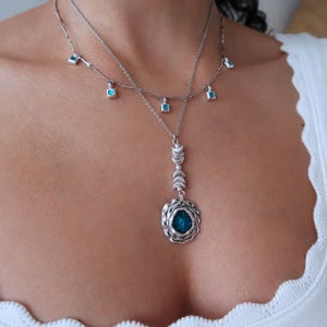 May include: A silver necklace featuring a turquoise pendant and several smaller turquoise charms. The pendant is a round, textured design with a turquoise stone at its centre. The necklace is layered with a delicate chain adorned with square turquoise charms.