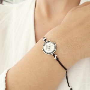 Pode incluir: Uma pulseira prateada com um pendente circular. O pendente tem uma superfície texturizada e uma pequena gema central. A pulseira é enfiada num cordão preto com nós deslizantes ajustáveis e pequenas contas prateadas.