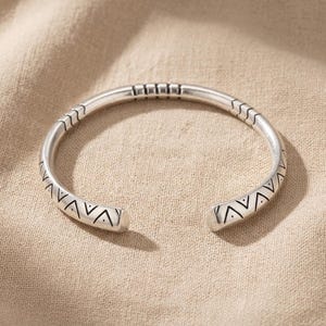 Op de afbeelding: Een zilverkleurige open armband met een gepolijste metalen afwerking. De band is versierd met gegraveerde geometrische patronen, waaronder herhalende driehoeken met centrale stippen en lineaire chevron-achtige markeringen op het oppervlak.