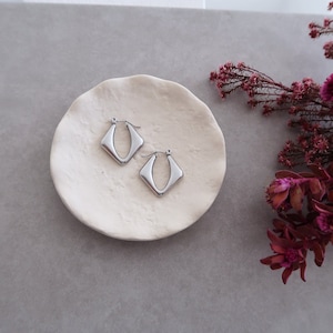 Op de afbeelding: Een paar zilveren creolen met een geometrisch, ruitvormig ontwerp. De oorbellen worden gepresenteerd op een wit keramiek schoteltje.