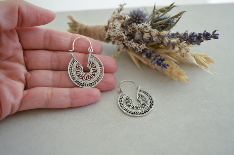 Puede incluir: Un par de pendientes de aro plateados con un dise&ntilde;o calado. Los pendientes tienen forma circular con un patr&oacute;n decorativo con un centro en forma de sol y detalles intrincados en los bordes. Se muestran sobre una superficie gris claro.